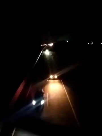 This media file is a short video clip recorded at night, likely from the perspective of a vehicle driving on a road. The primary subject is the road itself, illuminated by the headlights of the vehicle and a distant street light. The scene is dark, typical of nighttime. There are no visible people.

The activity taking place is driving. The perspective suggests the camera is mounted within or on a moving vehicle, facing forward. The road appears to be paved, with lane markings visible, indicating it's a highway or a well-maintained road. The presence of lane markings and the distant street light suggest an urban or suburban setting, or at least a developed area with infrastructure.

Notable details include the time of day, which is clearly night. The weather is not explicitly indicated, but the clear visibility of the road and lights suggests no significant precipitation or fog. There are no explicit location cues in terms of landmarks or signs, other than the implied road infrastructure. The mood is calm and solitary, as there are no other vehicles or people immediately apparent. The bright headlights cut through the darkness, creating a sense of forward motion and focus on the path ahead. There is no visible text in the media file. The general setting is a dark road at night.