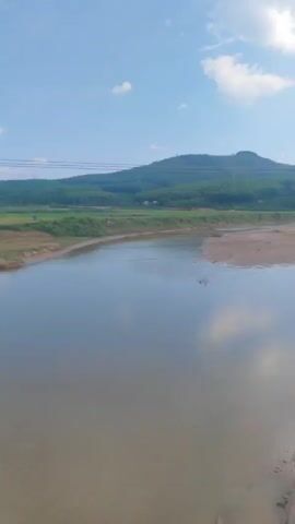 The video shows a moving view from a train, passing over a river and across an expanse of farmland under a partly cloudy sky. The water is a murky brown, reflecting the sky, and the landscape includes patches of green and dry, brown fields. Power lines stretch across the sky, supported by white utility poles. There are low hills on the horizon. The train ride seems smooth despite the shaky camera.