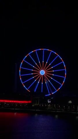 The video shows a Ferris wheel at night. The Ferris wheel is lit up with blue and red lights that change to blue and green lights. The Ferris wheel is located near a body of water, and there are buildings visible in the background. The Ferris wheel is slowly rotating.