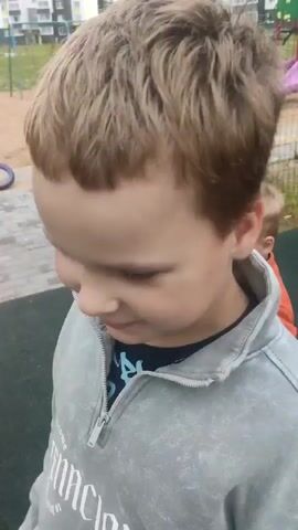 A young boy in an orange shirt and green pants stands on a paved surface, rolling up the sleeves of his shirt. He looks to his left. In the background, two bicycles are parked on a dirt area. A fence separates the paved area from the dirt area and a building complex. Another boy in a grey hooded jacket walks past in the foreground, obscuring the camera's view of the first boy's face. A second later, the boy in the orange shirt is visible from the side, looking to his right. A third boy in a t-shirt and patterned shorts is in the background, holding a basketball and looking at the boy in the orange shirt.
