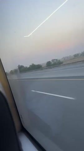 The video captures a view from inside a moving vehicle, likely a car or bus, on a multi-lane highway. The perspective is through a side window, showing the road, a concrete barrier, and guard rails to the right. Beyond the highway, a rural landscape with sparse trees, bushes, and fields stretches into the distance.

The sky exhibits soft, diffused light, with a faint pinkish-orange hue near the horizon at 0:00, suggesting either early morning (sunrise) or late evening (sunset). The overall light is gentle, indicative of dawn or dusk, possibly with some atmospheric haze. The vehicle appears to be moving at a moderate to high speed, evidenced by the motion blur, particularly on the road and immediate surroundings.

There are no people visibly present in the frame. The interior of the vehicle is minimally visible in the bottom left corner. No text is visible within the video itself. The background audio contains spoken words in what sounds like Punjabi or Urdu, further suggesting a location in Pakistan, consistent with the provided context of Thatta Haji.