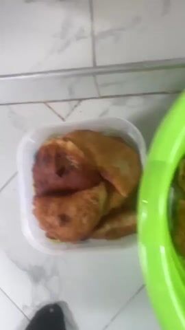 The video shows a person's hand reaching into a green plastic bowl. The bowl contains several fried food items that appear to be golden brown and some appear to be split open, revealing a filling. The food items are unevenly shaped and some are partially submerged in a clear liquid. A white plastic container with colored polka dots is visible in the foreground, partially obscuring the view of the bowl. The background appears to be a tiled floor. The lighting is dim.