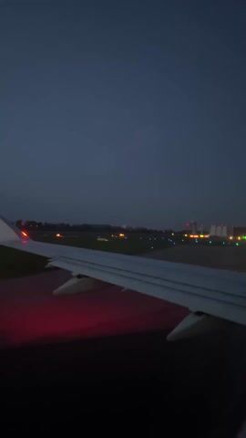 From the interior of an airplane, a view of the wing with a dark blue sky visible in the background.  The plane is on a runway, visible due to red and green lights.  Further down the runway are building or tower-like structures with multiple lights.  The angle of the video is slightly tilted.
