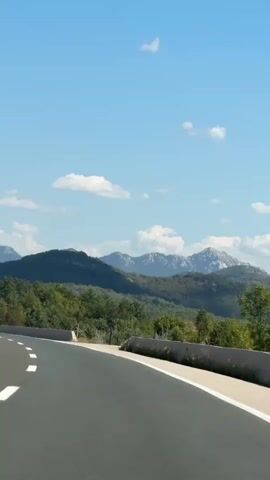 The video shows a view from a moving vehicle on a highway. The road is asphalt and has white lane markings. To the right of the road, there is a concrete barrier and then a fence. Beyond the fence is a landscape of green hills and mountains under a bright blue sky with scattered white clouds. The vegetation appears to be dense forest and scrub. The mountains in the distance are rugged and have rocky peaks. The overall impression is of a scenic drive through a natural landscape on a clear day.