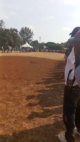The video shows a running race taking place on a dirt track.  Several runners are visible, sprinting towards the camera. Spectators are standing along the sidelines, cheering on the racers. In the background, there's a white tent and trees under a bright sky with light clouds.  The ground appears to be dry, with patches of brown grass.  A person with a red hat is partially visible in the foreground.