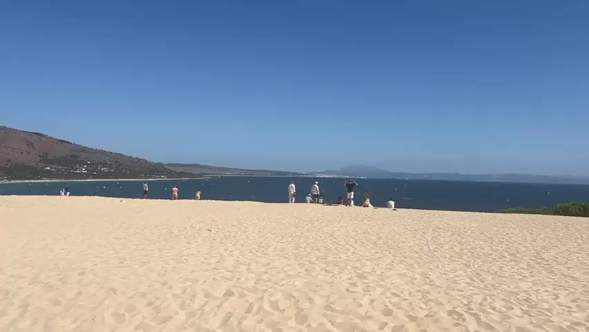 A wide shot captures a sandy beach with a clear blue sky overhead. In the middle ground, a group of people, some standing and some sitting, are visible near the edge of the sand dunes. The sea stretches out behind them, with a distant coastline and mountains on the horizon. The overall impression is a peaceful, sunny day at the beach.