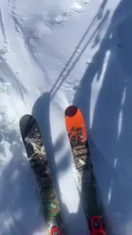 The video opens with a shadow of a person and their ski equipment on a snowy surface. The camera then pans up to reveal a snow-covered landscape with low-lying evergreen trees and a large, snow-covered mountain in the background under a clear blue sky. The scene is bright and appears to be filmed during the daytime.