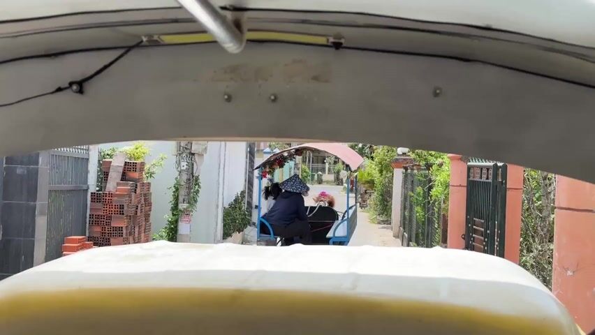 The view from the inside of a motorized vehicle, perhaps a tuk-tuk or similar vehicle, shows the back of a blue and red covered three-wheeled bicycle cart with two occupants, an adult wearing a blue long-sleeve shirt and a hat, and a child, riding along a narrow, walled road in a residential area. The vehicle that houses the camera is light yellow and the view out is framed by the vehicle's roof and pillars. On either side of the road are fences, houses, and greenery. Some bricks are visible stacked on the left side. The sky is not visible.