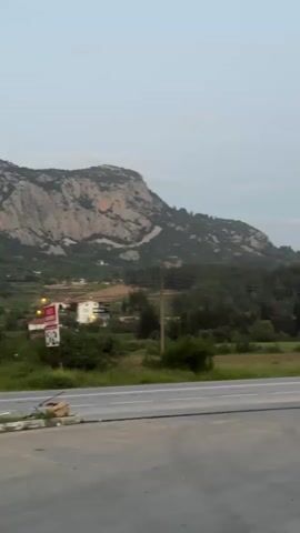The video captures a panoramic view of a rural landscape near Akseki, Turkey, during what appears to be dusk or dawn. The camera slowly pans from left to right, revealing a scenic vista dominated by mountains.

In the foreground, a paved road with two lanes is visible, stretching horizontally across the frame. To the left of the road, a white and red sign structure is partially visible, though its text is unreadable. Beyond the road, a strip of green grass and scattered bushes leads to a dense forest of trees.

The midground features several houses with light-colored facades and red-tiled roofs, nestled among the trees. Some of these houses have lights on inside. Power lines with visible poles run across this section of the landscape.

In the background, large, rocky mountains rise majestically. The mountains on the left are more sharply defined and show distinct rock formations and some tree cover, while the mountains further right appear hazier and more distant, fading into the soft, light blue-grey sky. A white tarp structure is visible in the lower right, beyond the road and trees. The overall impression is one of tranquility and natural beauty.
