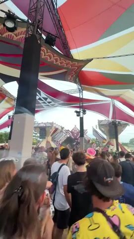 A low-angle, shaky, wide shot captures a crowded outdoor festival setting under a large, colorful, tent-like canopy. The canopy is a vibrant mix of red, yellow, white, and black geometric patterns, creating a festive atmosphere. A tall, black speaker tower is positioned in the foreground, obscuring part of the view. In the middle ground, a stage is visible with a complex, pyramid-like structure adorned with geometric patterns and a large, pale face. Numerous people, many with their backs to the camera, are densely packed, suggesting a concert or event. Some individuals are wearing hats, and one person in the foreground has a bright yellow and pink patterned shirt. The sky above is a pale blue, indicating it is daytime or early evening. The overall impression is of a lively, large-scale gathering.