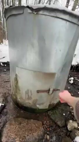 A person in a plaid jacket opens a metal door on a large, cylindrical metal tank. The tank is outdoors, surrounded by rocks, snow, and bare trees. A metal pipe is seen next to the tank. The person uses a metal dipper, attached to a chain, to scoop water out of the tank.