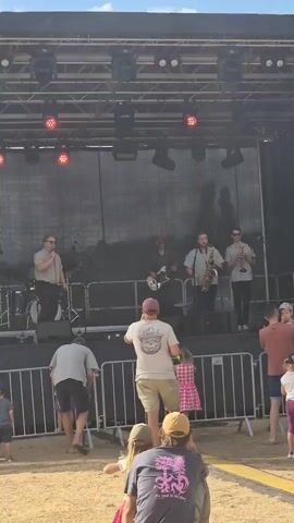 A band is performing on a raised stage outdoors. The band consists of a drummer on the left, a male singer in the center, a guitarist behind the singer, a male saxophonist to the right of the singer, and a trumpeter on the far right. The stage has a black backdrop and is lit by red stage lights. In front of the stage, a metal barricade separates the audience from the performers. Several people are visible in the foreground, including children and adults, watching the performance. The ground in front of the stage appears to be sandy or dirt. The sky is visible above the stage structure.