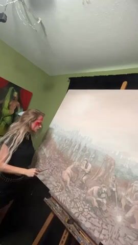 A woman with long blonde hair and red paint on her face is working on a large painting in a room with green walls. She is wearing a black top and skirt with a studded belt. The painting on the easel depicts a group of people and a dog in a dusty, war-torn landscape with a city in the background. The upper part of the canvas is still blank. Another painting hangs on the wall to the left, featuring a figure in green. The room is brightly lit. The woman is using a long brush to add details to the painting.