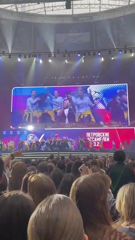 The video shows a stage performance in front of a large audience. The performers are dressed in various outfits, including tracksuits and casual wear. There is a large screen above the stage displaying close-ups of people in the audience, including women making eye contact with the camera, and the performers on stage. The stage is brightly lit with spotlights. The audience is mostly composed of young adults and teenagers, many of whom are holding up their phones to record the performance. The overall atmosphere is energetic and lively. There is text in a foreign language (possibly Russian) on the screen.