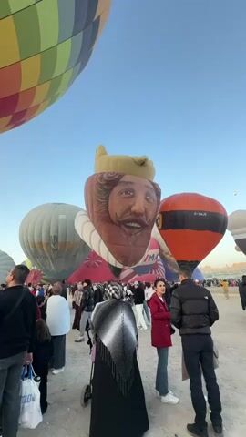 The video captures an outdoor gathering of people in what appears to be a desert or open field, with a clear blue sky overhead. The ground is dusty and uneven. The central focus of the scene is a collection of hot air balloons. Several are visible, including one large, colorful balloon with a geometric pattern dominating the upper left of the frame. Another prominent balloon features a large, stylized face of a man with a beard and a crown, resembling a cartoon king. An orange and black balloon is also clearly visible. A crowd of people, dressed in a variety of casual clothing, are milling around the area, some standing, some walking. A few individuals are captured in mid-action, such as a woman in a red jacket and jeans looking towards the camera, and a man in a black jacket and dark pants also facing forward. A person is pushing a stroller. The atmosphere seems relaxed and festive, with people observing the balloons. The lighting suggests it is daytime, possibly late afternoon given the angle of the sun.