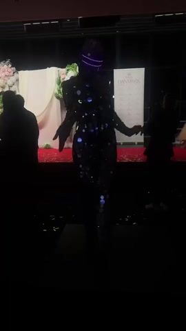 A person wearing a full bodysuit with round mirror-like decorations stands on a platform in a dimly lit room. The figure has a light-up visor on their head. In the background, other people in formal wear stand on a red carpet in front of a backdrop. The backdrop features white curtains and floral decorations. A "Miss Denmark" banner is visible on the right side of the backdrop. The setting appears to be a formal event, possibly a beauty pageant. There are several other individuals visible in the background in formal dresses and suits.