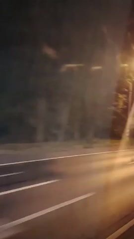 The video shows a blurry, nighttime view from a moving vehicle. The scene is illuminated by streetlights and the headlights of other vehicles, which create streaks of light across the road. Trees and some sort of poles line the side of the road. The road itself appears to be a multi-lane highway. The overall impression is one of speed and transit.