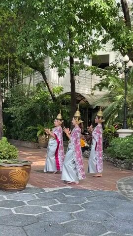 Three women wearing traditional Thai dance costumes are seen walking barefoot in a courtyard area. The women are dressed in silver, pink, and white outfits with elaborate headdresses. They are moving their hands in a stylized manner, suggesting they are performing a dance. The courtyard is paved with irregularly shaped stones, with greenery including trees, plants, and potted shrubs around. A large decorative pot with a rooster design sits in the foreground. A building with a white facade is visible in the background.
