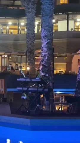 A man is playing a keyboard next to a pool at night. He is wearing a colorful shirt and is seated on a stool. There are palm trees behind him, and a building with lights on is visible in the background.