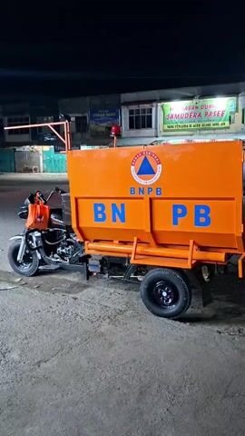 A still shot captures a custom-built vehicle, likely a motorcycle with an attached trailer, standing on a paved street at night. The vehicle is painted bright orange and has blue lettering. “BNPB” and “PB” are printed on the sides of the trailer, accompanied by a seal with “BADAN NASIONAL PENANGGULANGAN BENCANA” written around it. The trailer’s boxy shape suggests it's designed for hauling. The motorcycle has a dark color and an orange bag is hanging from its handlebars. A red emergency light is affixed to the back of the trailer, above the box. The background shows buildings with neon signs, one of which reads "SAMUDERA PASEE." The lighting is dim, indicating the scene is likely captured in a residential or commercial area at night. No people are visible in the frame.