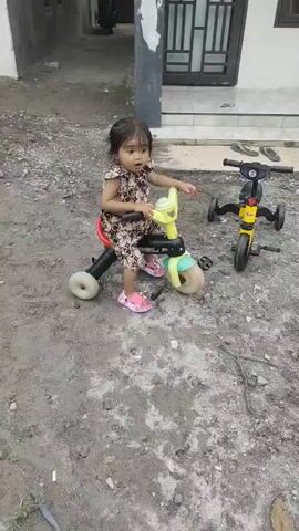 A young child wearing a floral-patterned dress and pink shoes sits on a yellow tricycle with a red seat. The tricycle has three wheels, two in the back and one in the front. The child is looking towards the camera with a pensive expression. To the right of the child, another yellow tricycle is parked on the ground. The ground is covered in gravel and dirt. In the background, a gray car is parked in front of a house. The house has a white facade and dark-colored doors and windows. Another child, wearing a blue shirt and pants, is visible in the distance. The scene appears to be outdoors, in what looks like a residential area. The video then shifts to a different scene where the same child, now wearing a blue shirt with "MALAYSIA" written on it, is holding a phone in her hand. She is standing on a gravel path, and a gray car is parked behind her. The video then returns to the initial scene of the child on the tricycle.