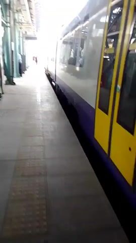 A train is stopped at a station platform. The train is white with a purple stripe near the bottom. The words HELLENIC TRAIN are visible on the side of the train. The platform is paved with textured tiles and there are pillars supporting an overhead structure. A sign with the word Exit is visible. Two people are walking away from the camera in the distance on the platform. The sun is shining brightly, creating a strong glare.