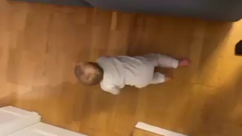 A baby wearing a white onesie is lying on its stomach on a wood floor. The baby is facing away from the camera and is in a crawling position but not actively crawling. The floor has a light brown wood grain pattern. Part of a dark gray object is visible at the top of the frame and a white object is visible at the bottom left. The baby's head is in the center of the frame and the baby is angled to the left.