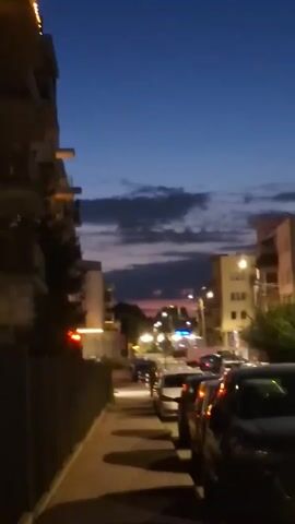 This is a vertical, shaky video taken from a sidewalk. On the left is a building, and on the right is a street lined with parked cars. The sky is dark blue with some clouds and a pinkish hue near the horizon, indicating it is dusk. Streetlights are on, illuminating the street and sidewalk. The cars appear to be dark-colored. The video is slightly blurry and lacks clear details about any specific people or actions.