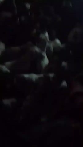 In a dimly lit scene, a cluster of mostly white goats are packed together. A light source from above illuminates the goats somewhat. The video seems to have been taken from inside a vehicle. It is unclear what is happening. There is an indistinct sound of someone talking.