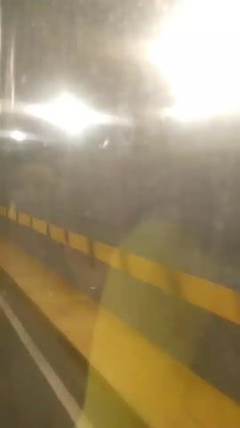 The video displays a view from inside a moving vehicle, likely a bus or train, looking out through a window. Bright lights are prominently reflected on the window's surface, suggesting either interior lighting or external streetlights/tunnel lights against a dark background, indicating it is possibly nighttime or the vehicle is in an enclosed space like a tunnel. Through the window, horizontal yellow stripes are visible, possibly part of a barrier, wall, or another passing structure. A faint, blurry reflection of a person wearing a light-colored (green or yellowish) shirt can be seen in the window. A loud, sustained horn sound, characteristic of a train or large vehicle, is audible around the 0:02-0:03 mark. No specific location cues, weather conditions, or visible text are present in the footage.
