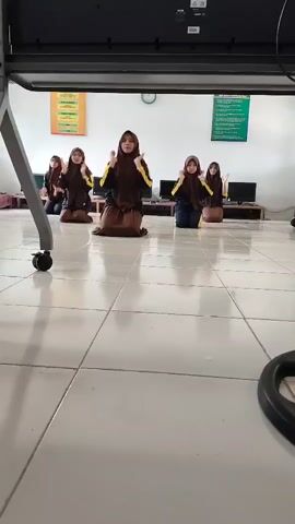 A low-angle shot captures five young women, dressed in brown and yellow Islamic hijabs and long skirts, kneeling on a tiled floor. They are positioned in a line, facing away from the camera, and are performing a synchronized dance. In front of them, a large black object, possibly a projector screen or a whiteboard, is visible. The room appears to be a classroom, with computers and posters on the walls in the background. The women move their arms in unison, demonstrating fluid and graceful choreography.