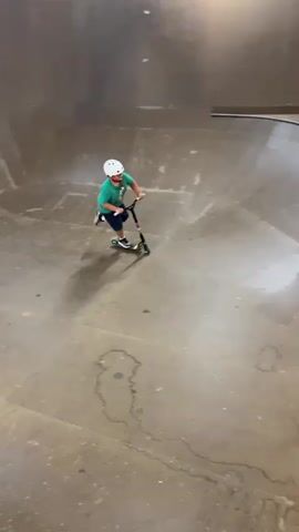 A young person wearing a green shirt, blue shorts, and a white helmet is riding a scooter in a skatepark. The skatepark appears to be made of concrete and wood. Another person is visible in the background. The young person on the scooter falls off, and the scooter lands on its side.