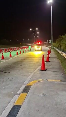 An overhead shot shows a highway at night. Orange traffic cones and red and white barriers are set up along the road, guiding traffic. A police car with flashing red and blue lights and bright headlights approaches the viewer. The car is moving towards the camera, and its lights illuminate the cones and barriers. The scene suggests a traffic stop or roadblock.