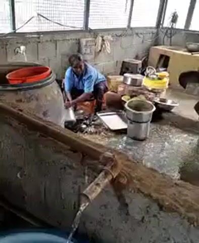 A person is sitting on a stool in what appears to be an outdoor or semi-enclosed kitchen area. They are wearing a blue shirt and a patterned lower garment. The person is actively washing or rinsing items from a large, dark, cylindrical container with an orange lid. In front of the person, on a surface, are various metal containers, bowls, and a tray, some of which appear to be clean and stacked, while others are in the process of being washed or are dirty. Water is flowing from a wooden pipe into a trough in the foreground, suggesting a water source and drainage system. The background shows a concrete wall with wire mesh or fencing at the top, and more cooking utensils and appliances, including a stove. The overall impression is of a functional, albeit somewhat rudimentary, food preparation or cleaning area.