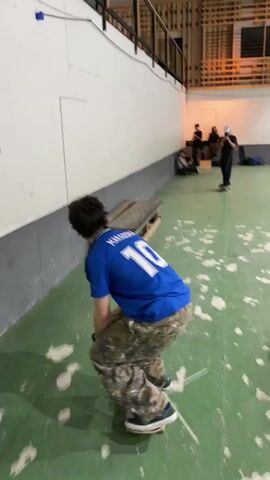 A male skateboarder in a blue shirt with the number 10 on the back and camouflage pants skates towards a wooden ramp in an indoor skatepark. He boards the ramp with his skateboard and rides up the side wall. Several other people are in the background, some sitting on a bench and others standing. The floor of the skatepark is green with white markings.