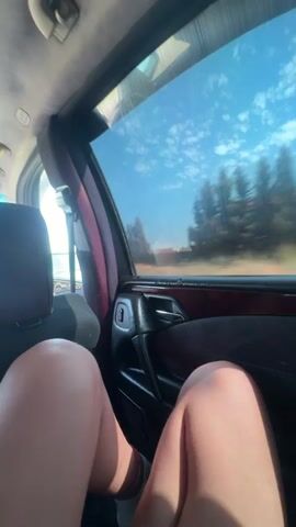 The video shows a first-person perspective from inside a car, looking out the passenger side window. The view outside is blurred due to the car's movement, showing a bright blue sky with scattered white clouds and trees. The interior of the car is visible in the foreground, including a person's legs and knees, the car door with an armrest and window controls, and the headrest of the seat in front. The lighting suggests it is daytime and sunny. The video appears to be a short clip, with the timestamp indicating approximately 4 seconds in length.