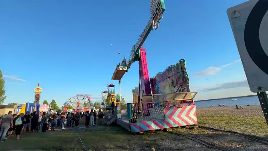 The video shows a busy outdoor carnival with several rides and attractions visible under a clear blue sky. In the foreground, a large, colorful amusement ride dominates the frame. It appears to be a thrill ride with a swinging arm and a platform that rotates and flips. The ride is adorned with vibrant pink, teal, and yellow stripes and various cartoonish graphics. To the left of this main ride, a queue of people stands waiting, suggesting the ride is in operation or about to begin. Further in the background, other carnival rides are visible, including a Ferris wheel with brightly colored cars and a tall, narrow tower ride with flashing lights. The setting appears to be a waterfront area, with a beach and the calm water of a lake or ocean visible in the distance. Several people are scattered on the beach, some sitting and relaxing. A sign with a large black circle is present on the right side of the frame, possibly indicating a game or target area. The overall atmosphere is lively and festive, characteristic of a summer carnival event.