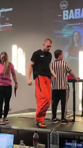 A male arm wrestler in orange pants and a black t-shirt walks onto a stage. He is accompanied by two referees in striped shirts. A large screen in the background displays "BABAEV'S CUP" and event details. The scene appears to be an arm wrestling competition.