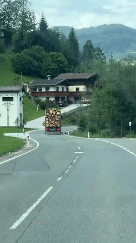 A logging truck carrying a load of logs drives down a winding road. The truck is in motion and appears to be turning a corner. The road is paved and has white lane markings. On the right side of the road, there is a slope covered in grass and trees. On the left side of the road, there is a building and a steep, grassy slope with trees. In the background, there are houses and more trees on a hillside. The sky is overcast.