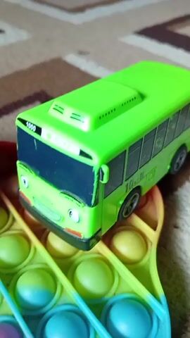 A close-up, slightly shaky video shows two toy buses, one green and one blue, positioned on a rainbow-colored pop-it toy. The green bus is larger and positioned closer to the camera, facing right. The blue bus is smaller and to the left of the green bus, also facing right. The pop-it toy is heart-shaped and has rows of colorful bubbles. The background is a carpet with a geometric pattern. The lighting is artificial.