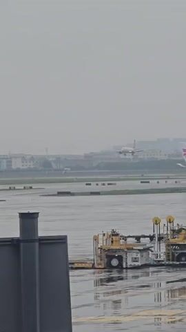 An airplane is approaching a runway on a hazy day. In the foreground, there is a grey, rectangular structure. Further back, a yellow and white vehicle with wheels is parked on a wet tarmac. The runway is also wet and reflects the dim light. Buildings can be seen in the distance.