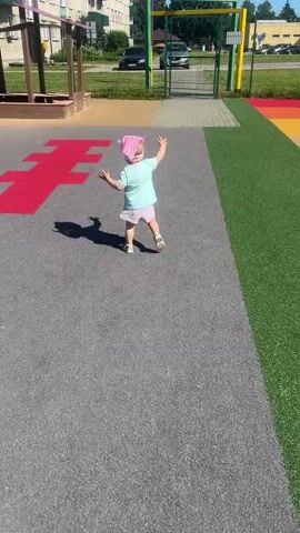 A young child wearing a pink hat and a light blue shirt with pink shorts is playing on a playground. The child is on a grey surface with red markings. The child is initially standing and appears to be dancing or playing, with arms outstretched. The child then crouches down and appears to be interacting with the red marking on the ground. In the background, there is a building, a fence, and a street with cars. The sun is shining, casting shadows on the ground.
