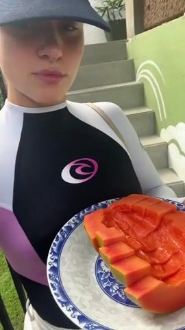 A person wearing a dark blue baseball cap and a long-sleeved white and black rash guard with a pink trim and a circular pink logo is holding a plate of sliced papaya. The person has a nose ring and is looking at the camera with a slight smile. The background shows a set of stairs and a green wall with a white wave-like design. The person appears to be outdoors, possibly in a tropical location, as indicated by the lush greenery visible to the left. The plate of papaya is cut in half and the slices are arranged in a circular pattern. The person's right arm is bent at the elbow, holding the plate steady. The lighting suggests it is daytime.
