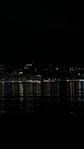 The video shows a nighttime view of a city skyline across a body of water. The city lights are reflected on the dark water's surface. The sky is completely black, indicating it is nighttime. There are no people or distinct objects visible in the foreground. The scene is peaceful and tranquil.