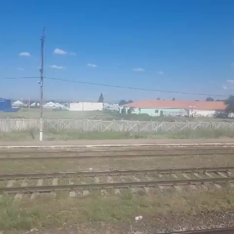 The video shows a landscape with train tracks in the foreground. Beyond the tracks, there is a white fence, some buildings, and trees. The sky is blue with a few white clouds. The perspective suggests the video is being filmed from a moving train. There are no people visible in the frame. The buildings appear to be a mix of residential and possibly industrial structures. The environment appears to be rural or semi-rural.