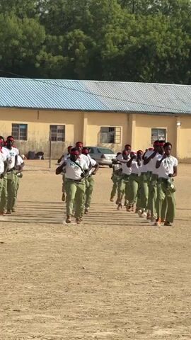 A group of around 15 people, possibly young adults, are marching in formation on a dry, sandy field. They are wearing matching green and white uniforms with red headbands and brown shoes. The uniforms have "NYSC" printed on the front. They are raising their arms in unison in what appears to be a military drill. Behind them is a yellow, single-story building with a blue and white striped roof. A car is parked to the right of the building, and green trees can be seen in the background. The sky is overcast.