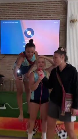Three people are in front of a large screen, which displays song lyrics. The person in the center is a young woman with blonde hair, wearing a dark blue and teal tank top and black shorts. She is holding a red microphone and singing with her eyes closed. To her left, a woman with dark hair pulled back in a bun, wearing a similar dark blue and teal tank top and grey shorts, is leaning down and laughing. She is holding a smartphone in her right hand, its flashlight on and directed towards the center. To the right of the central singer is a person with their hair tied back, wearing a black long-sleeved shirt and grey shorts. They have their left hand on the central singer's shoulder and are also singing. The background consists of a brick wall and a portion of a decorative light fixture. The floor appears to be covered in green turf.