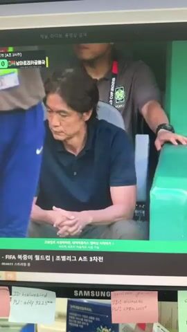 A medium shot shows a man sitting on a bench on the left side of the frame, with his arms resting on his lap. He is wearing a dark blue polo shirt. Behind him, a person wearing a grey shirt with a FIFA World Cup 2026 logo is standing and looking down. The background is out of focus. Below the screen, there are sticky notes and part of a Samsung monitor is visible. The screen displays a soccer match broadcast with Korean text.