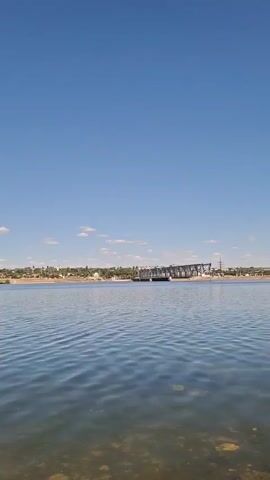 The video is an outdoor shot of a wide, calm body of water with gentle ripples on the surface. The sky above is a clear, vibrant blue with a few scattered, wispy white clouds. In the distance, across the water, is a long, flat horizon line. Along this horizon, there appears to be a shoreline with a low-lying settlement or town, characterized by scattered buildings and trees. To the far right of the distant shore, a large, dark structure, possibly a bridge or a dam, is visible spanning across the water. The foreground of the video shows the shallow, sandy or pebbly bottom of the water, with some submerged rocks or debris visible beneath the clear surface. The overall impression is a serene, sunny day by a large lake or river. There are no people or obvious actions depicted in the video.