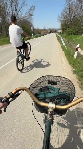 A first-person point-of-view video shows a person riding a bicycle on a paved road on a sunny day. Ahead of them, another person rides a bicycle while performing a wheelie. The first person's bicycle has a metal basket on the front with a dark green item inside. The road has a solid white line dividing the lanes and a metal guardrail on the right side. Grass grows alongside the guardrail. Trees are visible on both sides of the road, some still bare from winter. As the cyclists approach, a sign that reads "Джулюница Dzhulyunitsa" in Cyrillic and English comes into view, marking the entrance to a town. The scene is bright and appears to be rural.